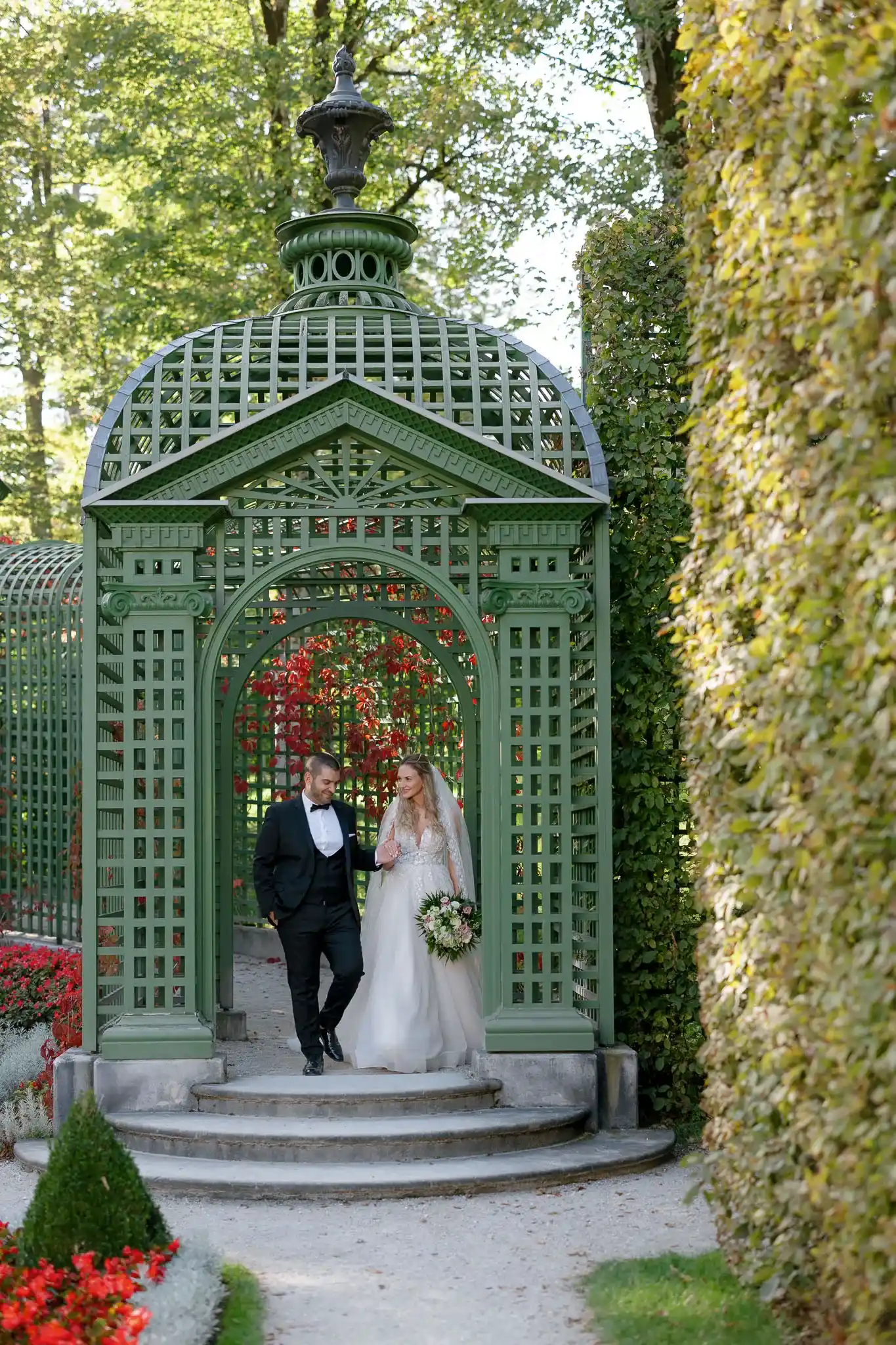 A smiling bride and groom walk under a green, ornate garden archway. The bride holds a bouquet, surrounded by lush foliage and vibrant flowers, evoking romance.