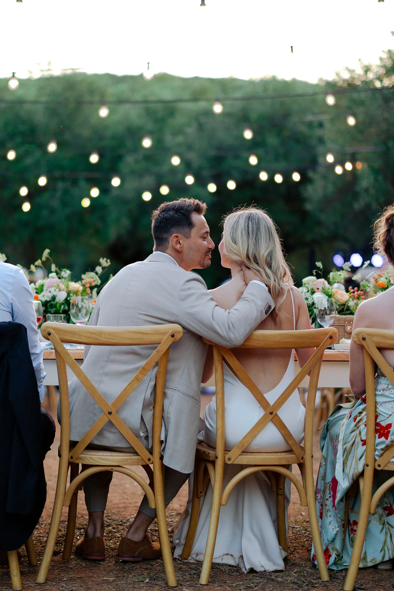 A romantic outdoor dinner setting with decorative string lights, featuring couples seated at a long table adorned with floral arrangements.