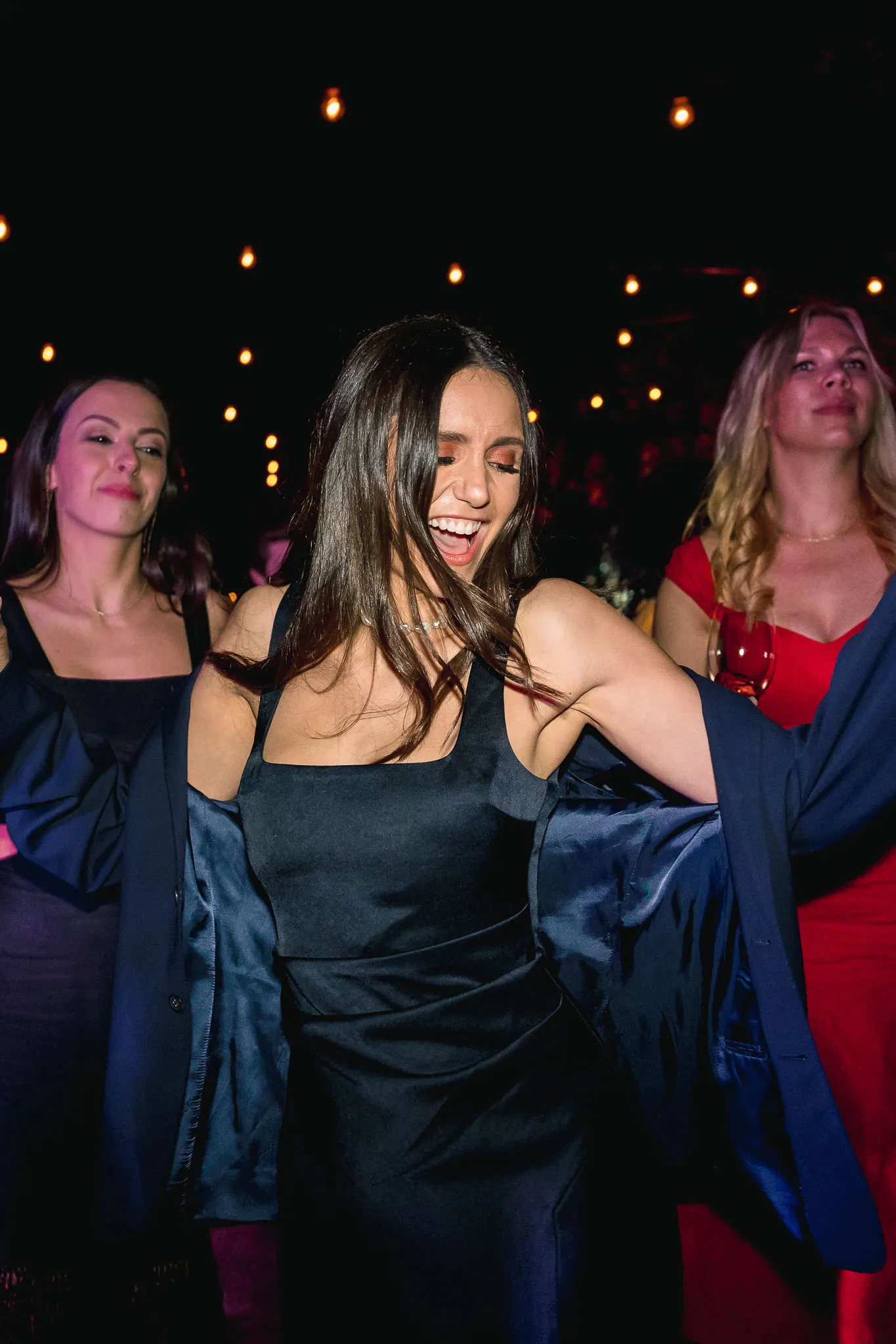 A woman joyfully dances with a wide smile, wearing a black dress and open jacket. Two women behind her are smiling under warm, dim lights. Festive mood.