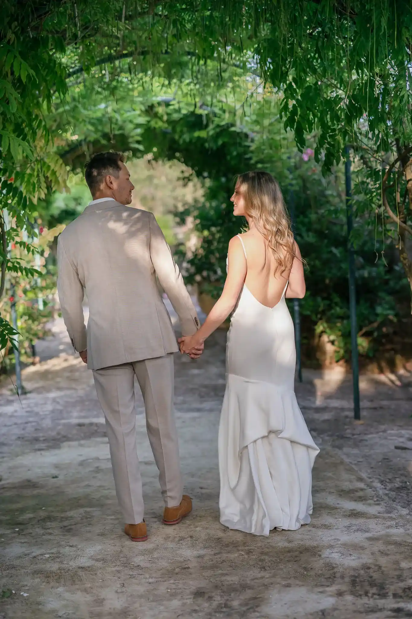 A couple holds hands while walking under a lush green archway. The woman wears a white, backless dress, and the man is in a beige suit. It conveys romance and serenity.