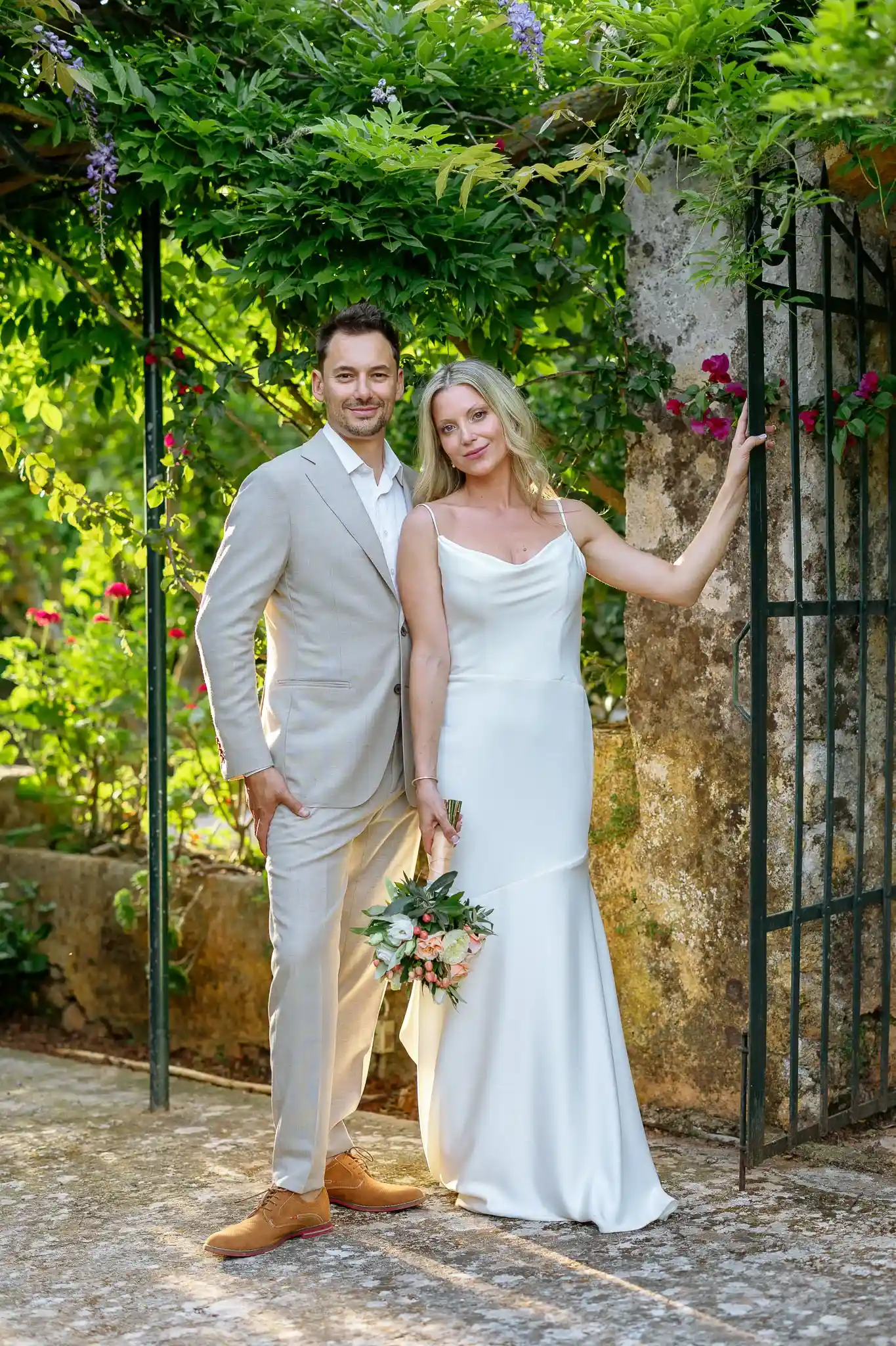 A couple poses in a garden under an archway of green foliage. The man in a light gray suit smiles, while the woman in a white dress holds a bouquet. Romantic and serene atmosphere.