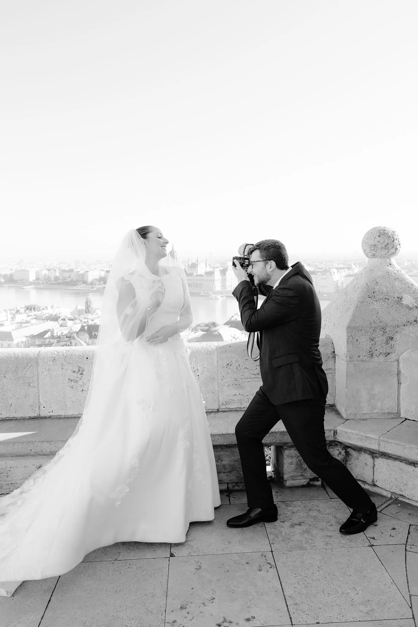 A candid black and white shot of the groom taking a picture of the bride with a professional camera on a scenic stone terrace overlooking the city.