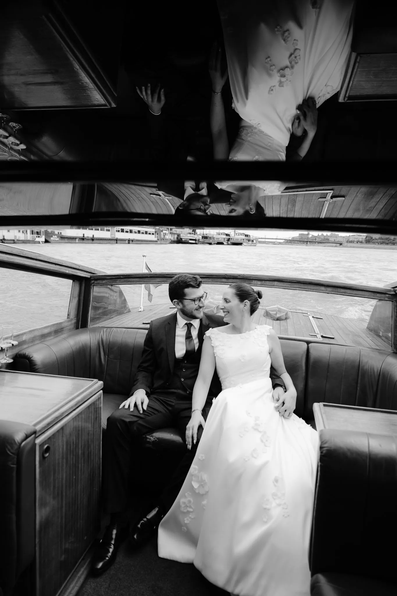 A couple in formal attire sits closely together in a boat, reflecting their love against a backdrop of water and distant buildings.